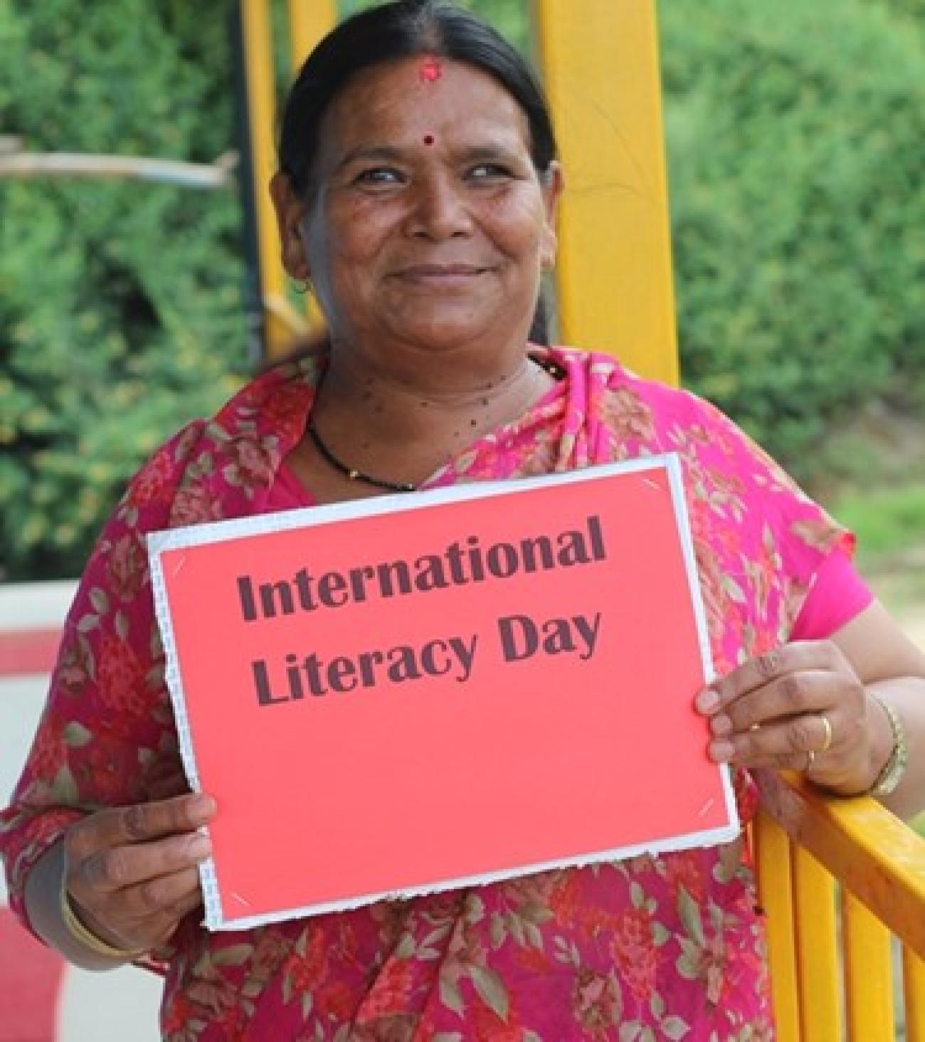 Chhalmaya wears a pink Saree with flower designs, holds a red paper that says International Literacy Day and smiles looking forward.