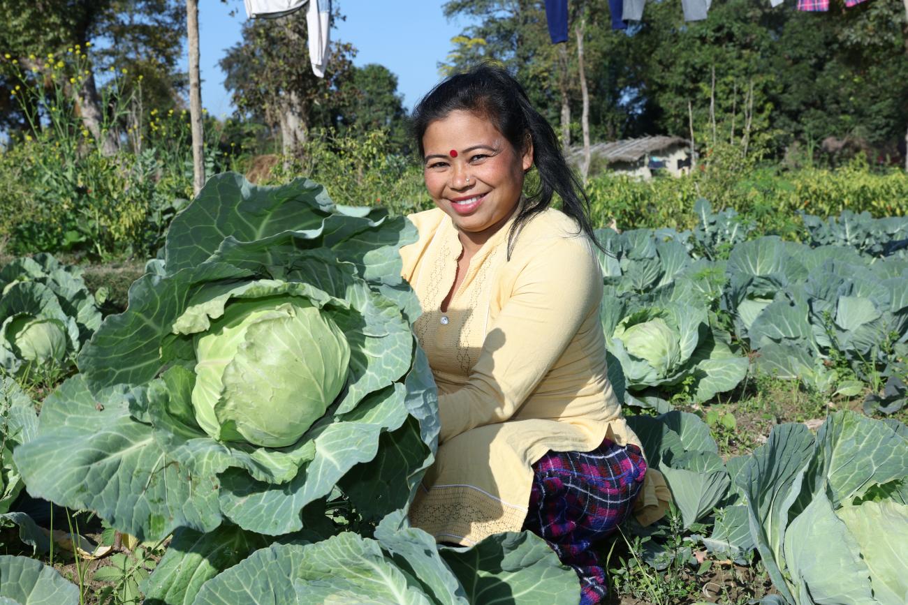 A woman stands in the field with a joyful expression, proudly holding a large cabbage she has harvested.