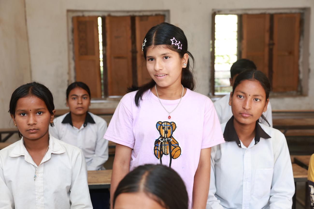 A young girl is standing in a classroom, surrounded by her friends who are seated on benches.