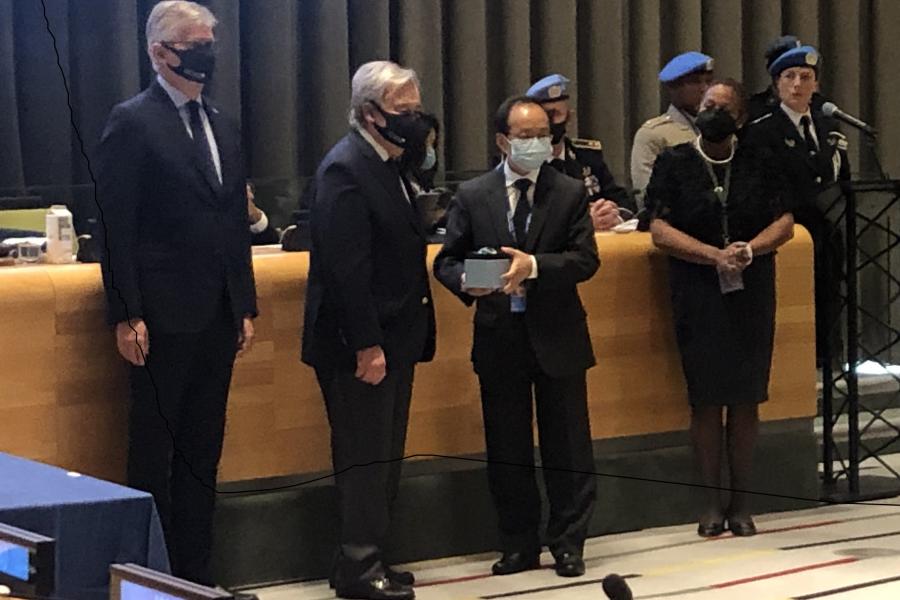 Four people stand beside each other, all formally dressed and wearing facemasks. The Secretary General stands in the middle, next to the Ambassador of Nepal who holds a square item in his hands.