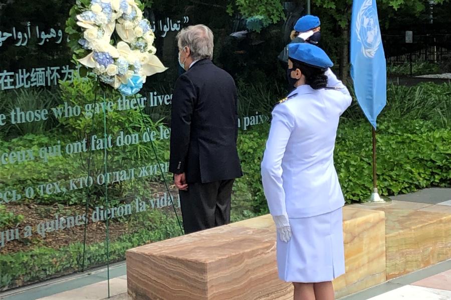 Secretary General stands in front of a wreath, back facing us. Female officer in uniform gives a salute a few meters behind him.