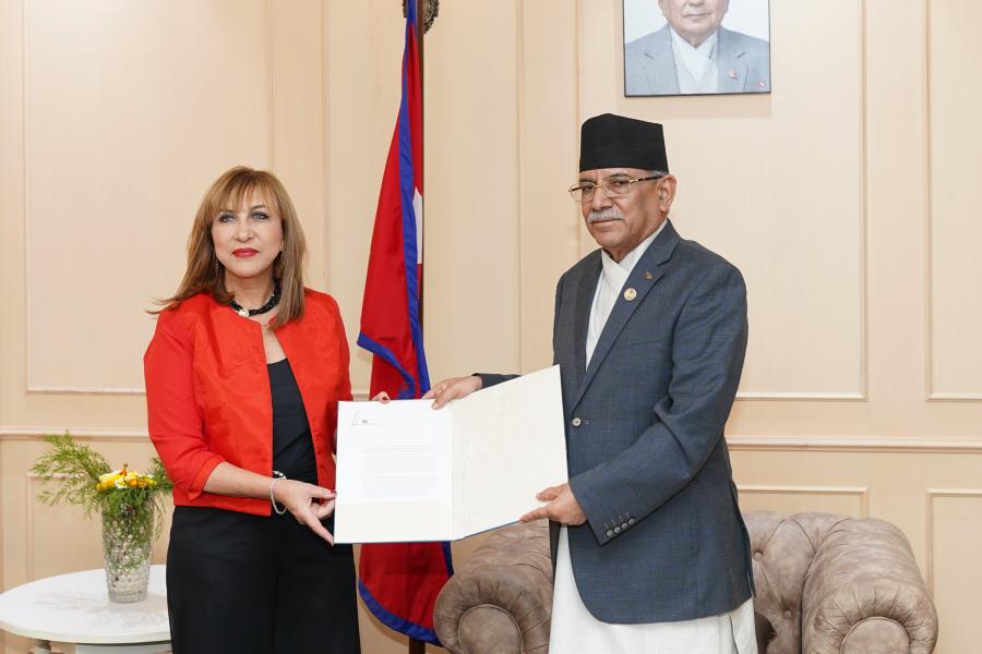 An image taken inside the Prime Minister’s Office in Singhadurbar. In the image the UN Resident Coordinator Hanaa Singer-Hamdy is presenting her credentials to Rt. Honorable Prime Minister of Nepal Pushpa Kamal Dahal. Resident Coordinator Singer-Hamdy is wearing a red coat and black pant. Rt Honorable Prime Minister is wearing Nepali national costume for men Daura Suruwal. 