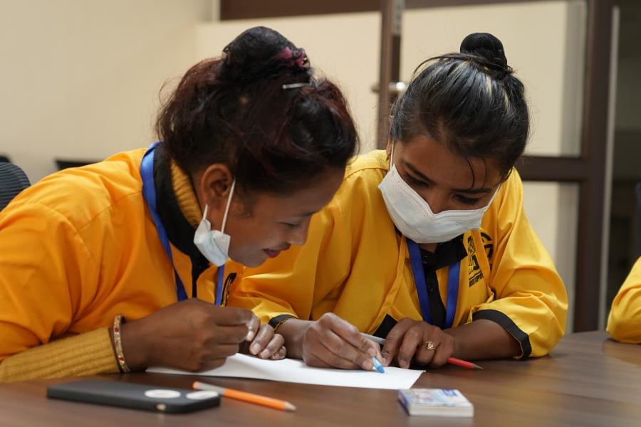 Two of the cleaning staffs wearing a yellow uniform are reading a paper on the table and highlighting important details on it.