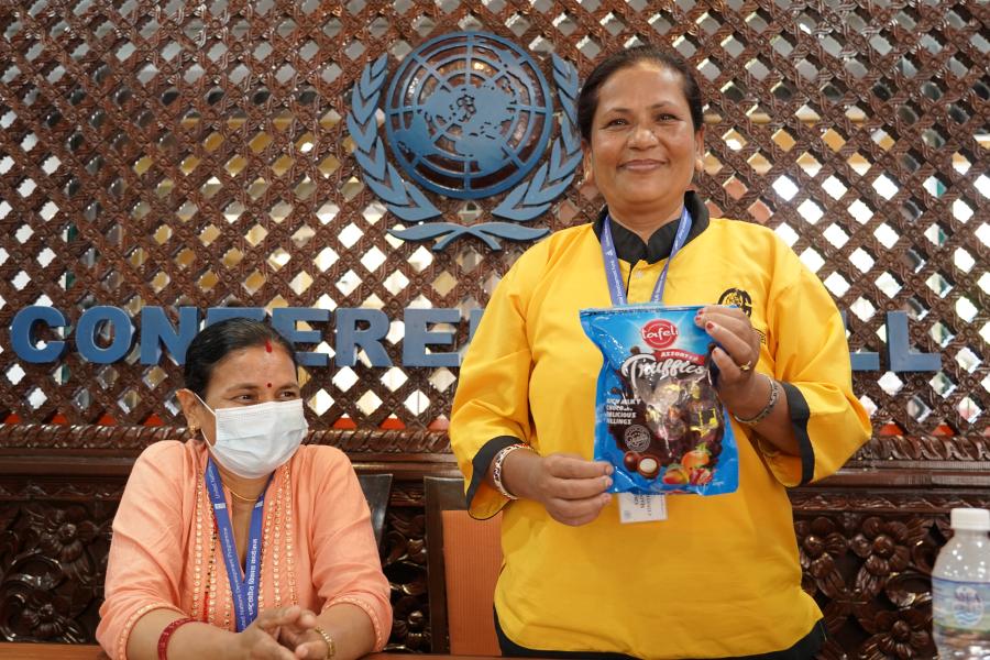 Sakuntala, one of the cleaning staff members is wearing her yellow uniform in the conference hall. She is standing and posing for a photo while holding a packet of chocolate truffles and smiling. the other lady wearing a baby pink colored kurtha in the picture is sitting down looking to the right.