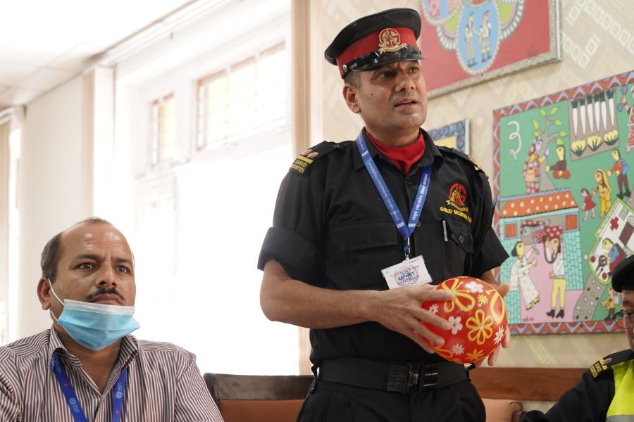 Karna Bahadur, one of the guards is wearing his black uniform, standing and holding a small red balloon with yellow flowered designs on it. There are two other people in the picture, sitting besides where Karna bahadur is standing and looking ahead.