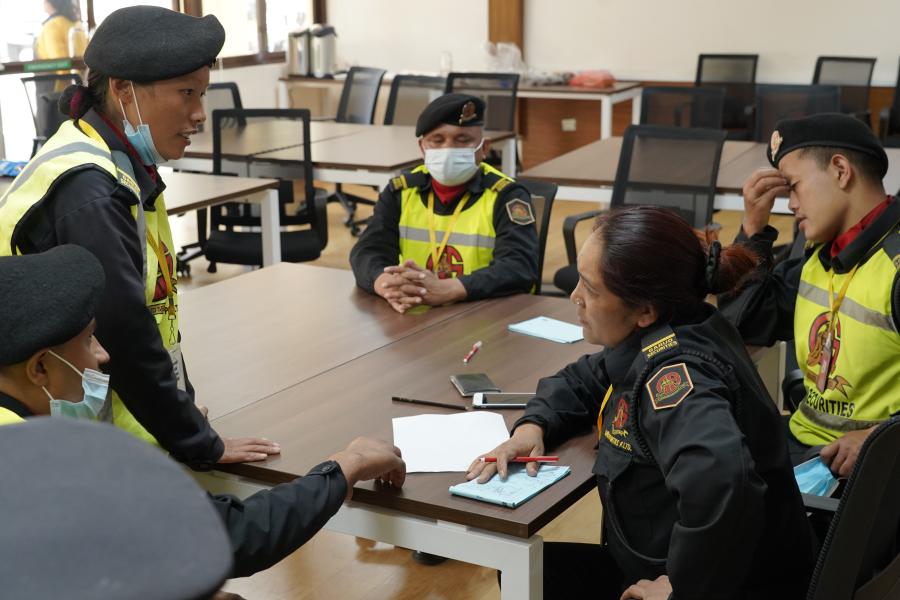 Five UN guards with their black uniform and neon vest are seen to be discussing. The lady guard has a pen in her hand and is taking notes of the discussion with her groupmates in the conference hall.