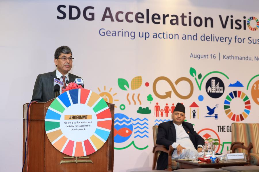 Dr. Min Bahadur stands behind the podium that has three mics on top. He is wearing a black suit and red tie. The podium has an SDG wheel on it.