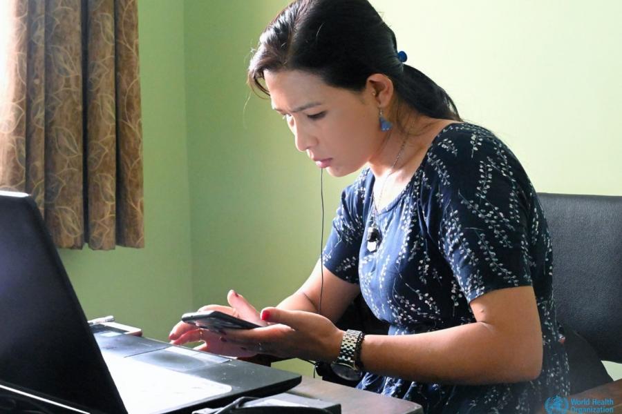 The counselor sitting on her desk in front of her laptop looks down intently at her mobile phone in her hand.