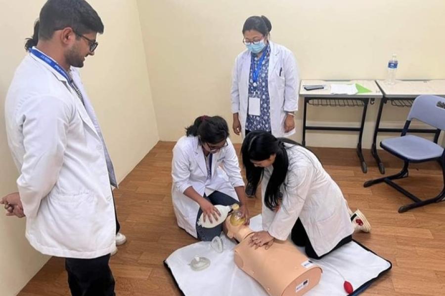 five men, four wearing white coats and the one in the middle wearing blue shirt demonstrates Advanced airway management with a dummy that's on the table.  A trainer demonstrating Advance Airway Management at the skill station. 