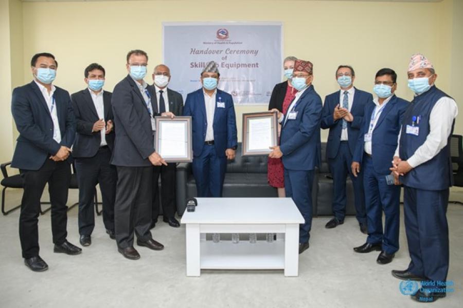 Ten men wearing suits are standing, with two of them holding certificates in the middle.  It is the signing of the MoU in Bir Hospital, Kathmandu