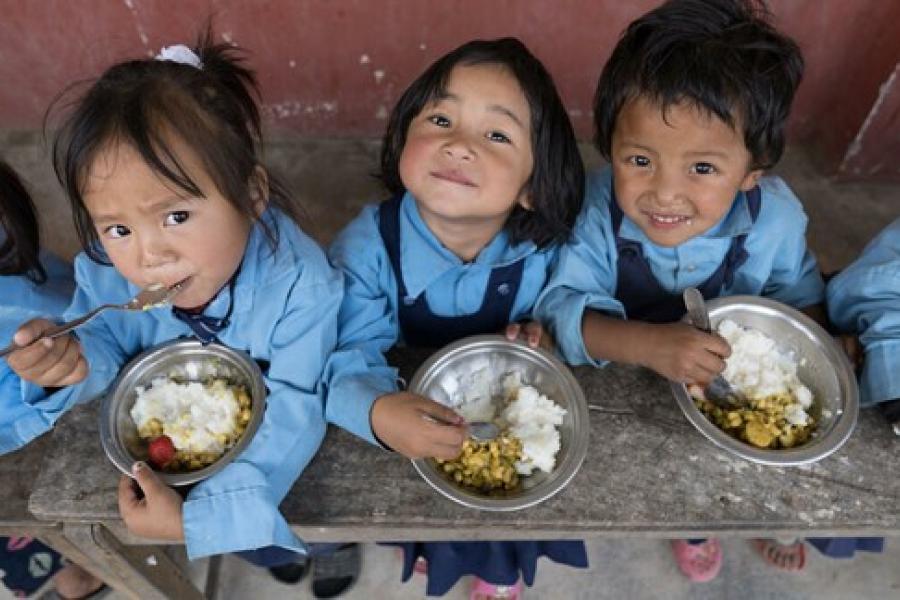 Three children sitting on a bench, looking above and smiling at the camera while enjoying their curry and rice with a smile. 