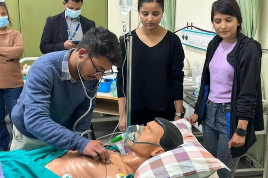 A dummy is laid in the bed, and a participant wearing a blue sweater uses his stethoscope to check on it. Two women behind the bed are looking at the participant. Participants practicing taking care of patients in ICU at the skill station.