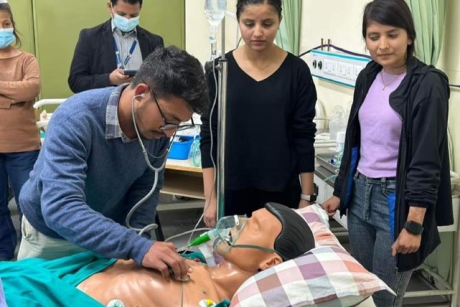 A dummy is laid in the bed, and a participant wearing a blue sweater uses his stethoscope to check on it. Two women behind the bed are looking at the participant. Participants practicing taking care of patients in ICU at the skill station. 