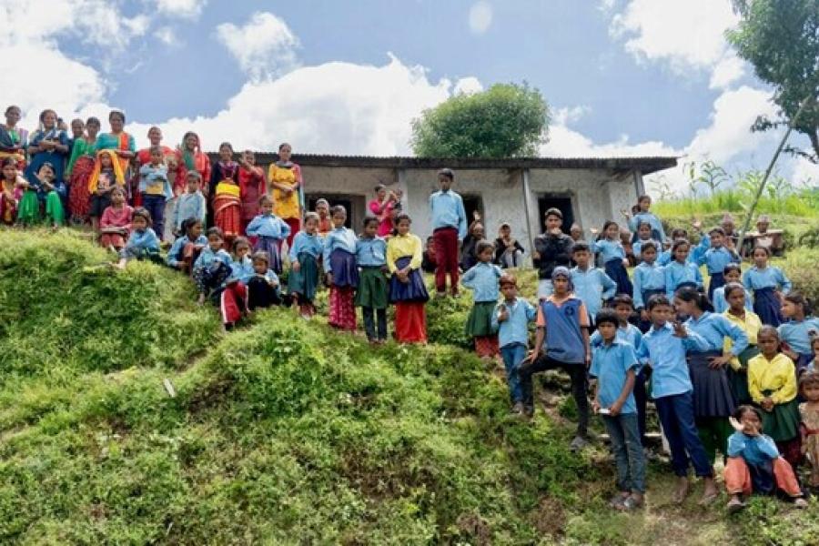 School children looking intently in front of them, some sitting while others standing as they watch the football match in an open area.