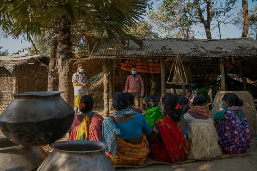 Women in vibrant saris sit clustered together outside wood and straw built shelters listening to a standing man and woman wearing masks. 
