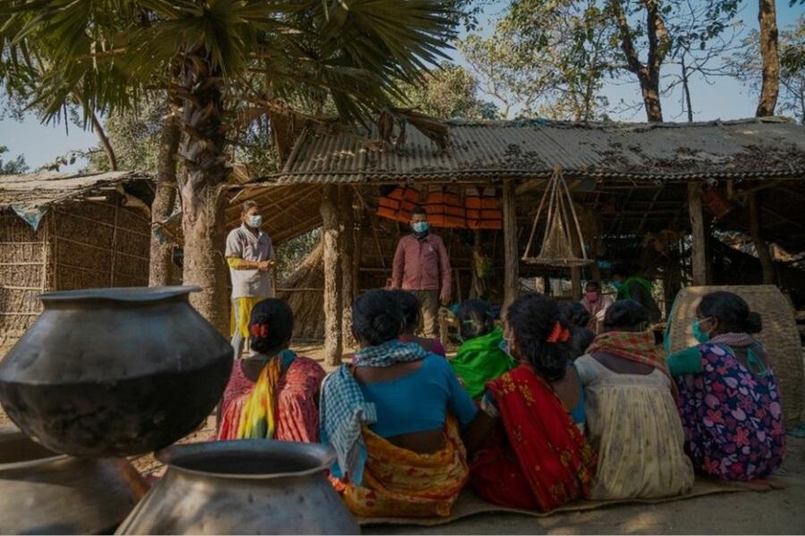 Women in vibrant saris sit clustered together outside wood and straw built shelters listening to a standing man and woman wearing masks.