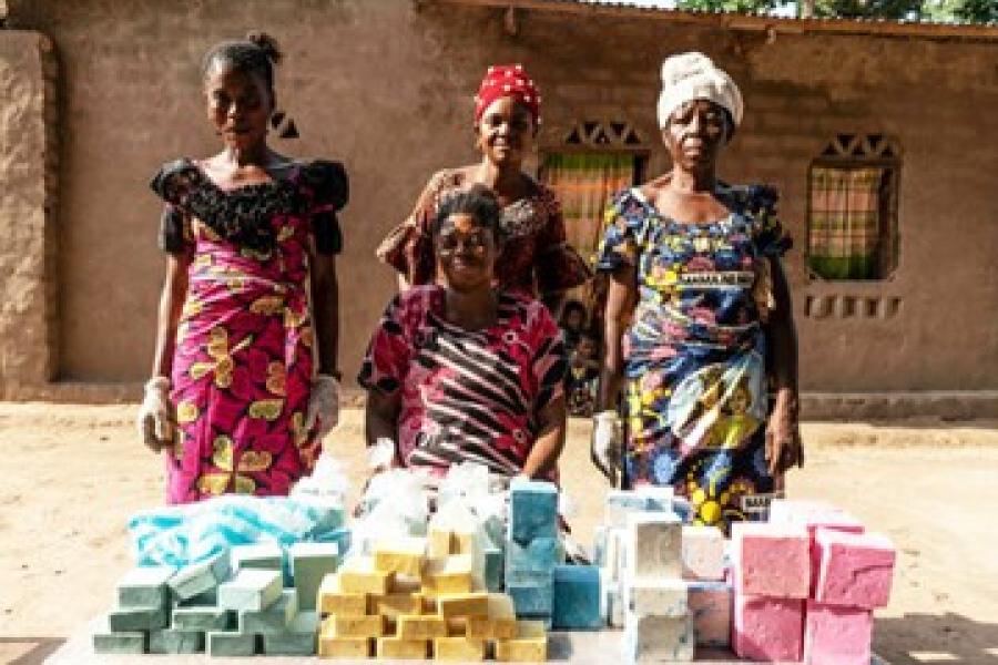 Four women in vibrant dresses, two of whom wear scarves tied to their heads, stand in front of a brick home next to a table of blue, yellow, white and pink soaps.