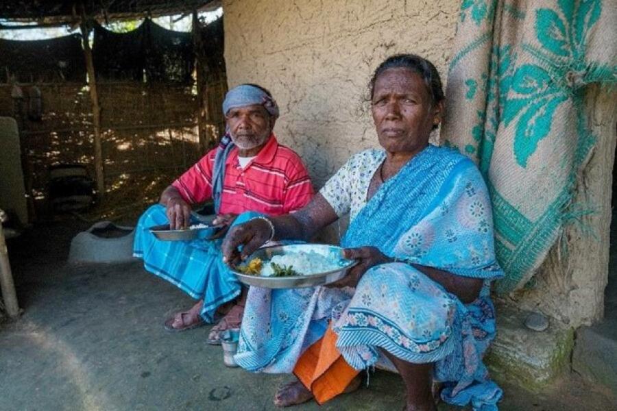 A women in a blue sari and man in a red shirt with a grey scarf tied around his head sit besides their home eating daal bhat.