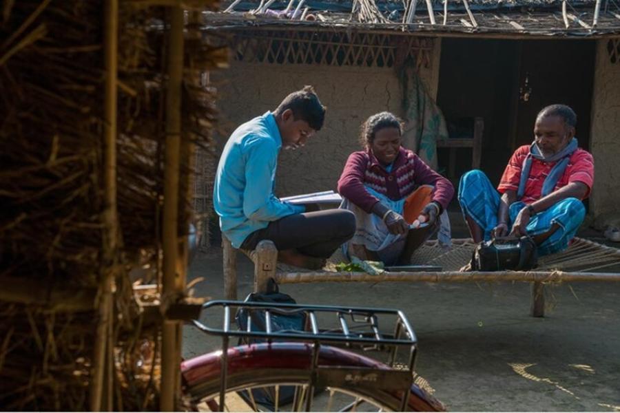 A man in a red shirt, a woman in a maroon sweater, and a boy in a blue shirt sit on a hammock. The man listens to the radio as the woman huts radish and the boy reads.