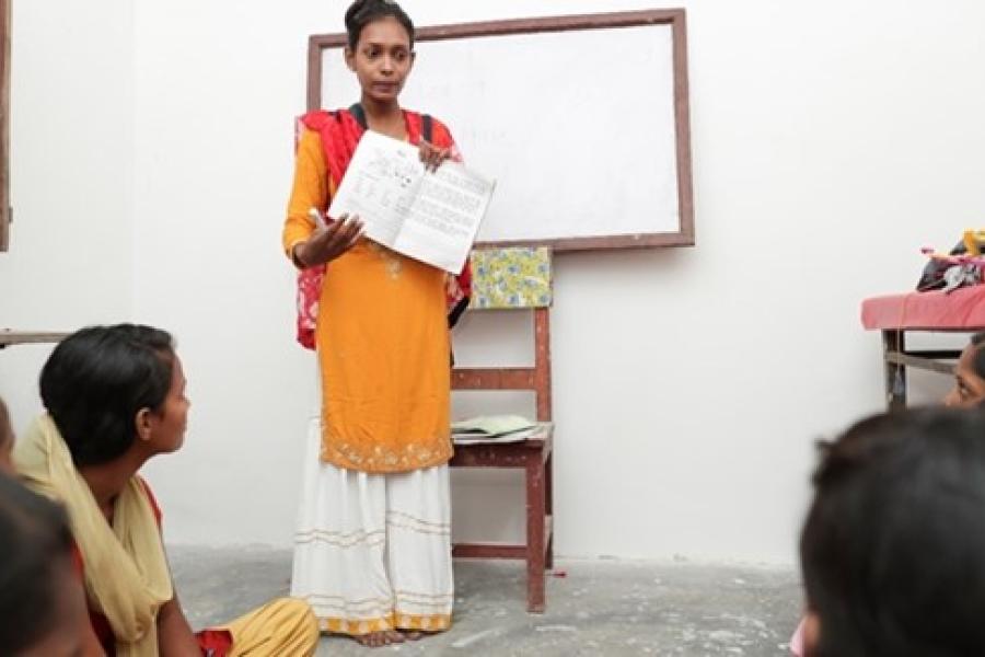  A woman wearing yellow kurtha and red shawl is standing in front of a whiteboard holding and showing the texts and pictures in the text book to the people sitting in front of her.