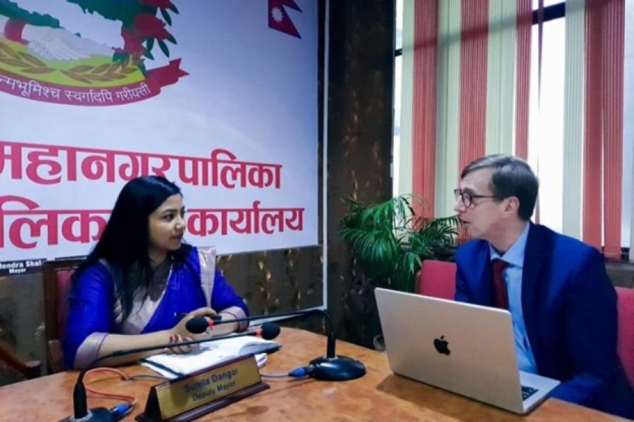 The SAFER Nepal Mission Team in A woman wearing a blue saree is discussing with a guy wearing a suit beside her. They are in from of a table, with a laptop, papers and a mic on top of it.