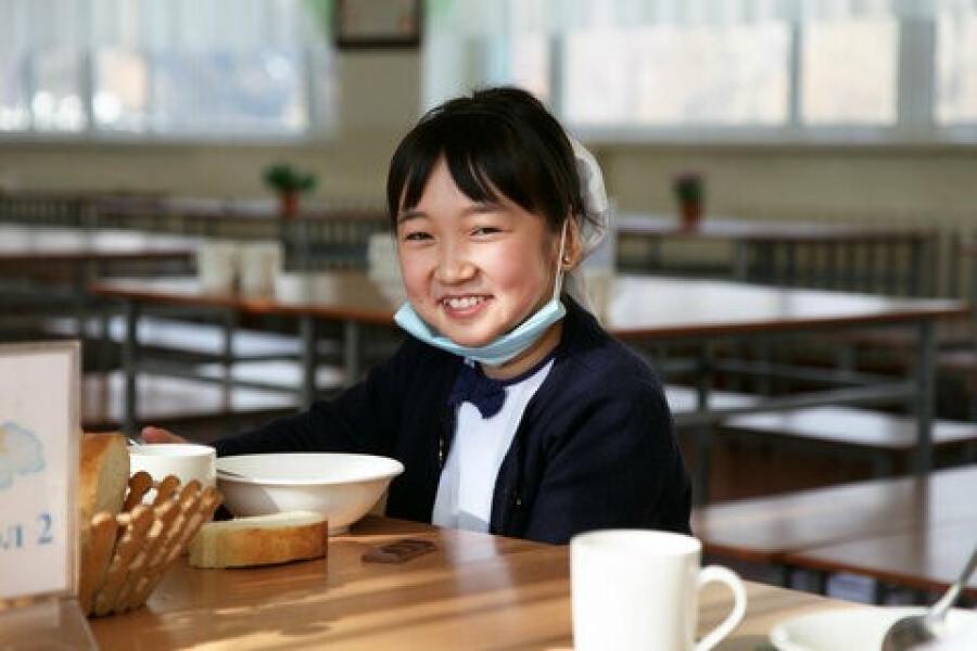 A girl sitting in the dining table as she holds a spoon. She has a blue mask below her chin and is smiling ahead.  Kyrgyzstan: School meals power one girl's dream of being a doctor