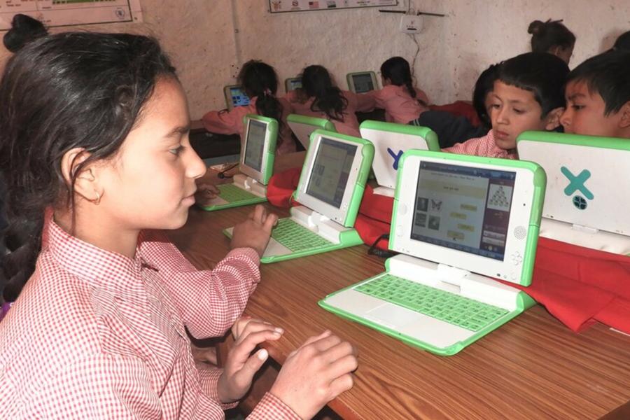 Children with red and white checked uniform sitting around the table in their classroom with their laptops open.
