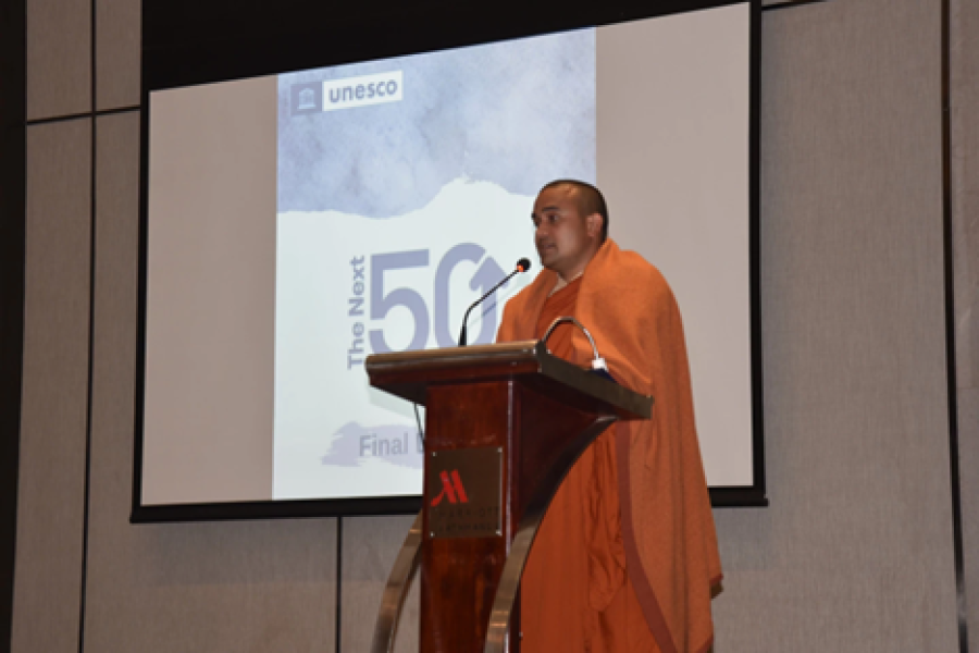 A man in an orange robe speaks into a microphone on stage next to a projected screen that labels 'the next 50'.