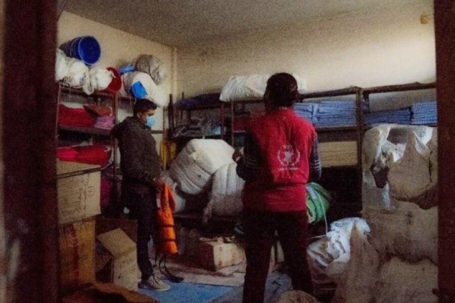 A man wearing brown clothes and a mask, and a women wearing a red WFP jacket stand in a room packed with boxes, lifejackets, tarpaulins, mattresses and sirens.