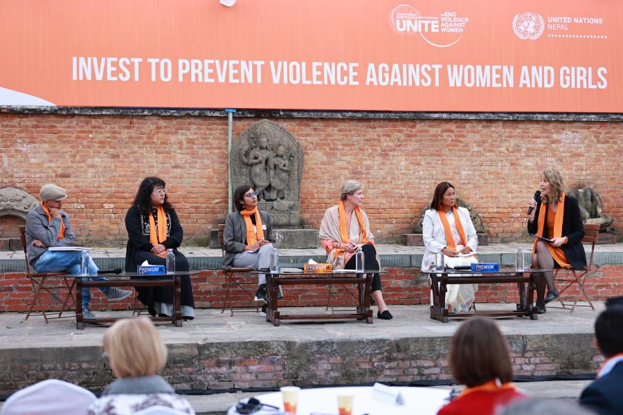 Five women and one man in orange scarves sit and discuss with a banner that says invest to prevent violence against women and girls in the background