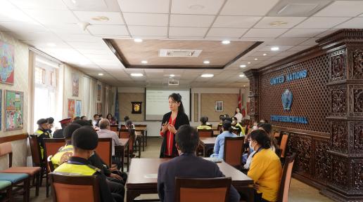 A women with red top and black cardigan is presenting slides to all the participants in the conference hall. There dais and didis wearing their respective uniforms and listening to the woman intently.