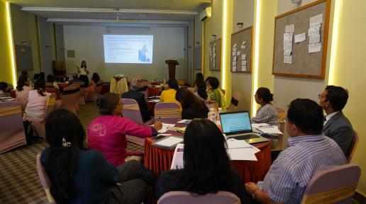 Lian Yong wearing a white top and black dress, holds a mic in her hand to commence the PSEA workshop for the attending participants in the room. Participants are viewing the projector screen, taking notes and listening to her intently while the opening session takes place.