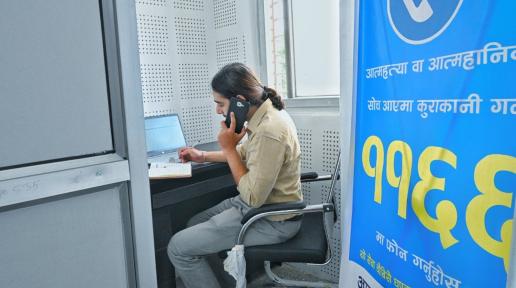 A man sits on his desk in front of the laptop inside his cubicle. He is holding his mobile and writing down on his notebook. 