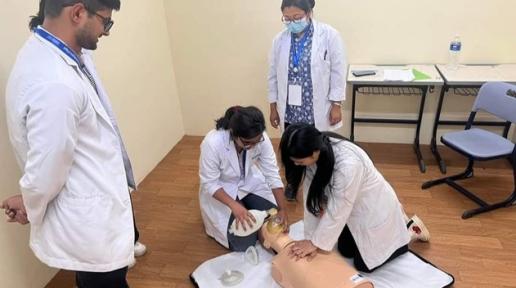 Two women with white coat practicing CPR on a dummy on the floor while a man and a third woman with white coat watch them inside a room.