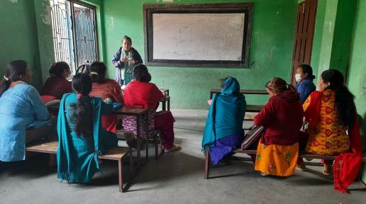 n a classroom setting, the mental health activist is standing and explaining to all the women sitting on benches with their backs faced to the camera. There are around nine participants listening to the activist.   mental health activist discusses with female community health volunteers on early identification of people in distress, 2021. 