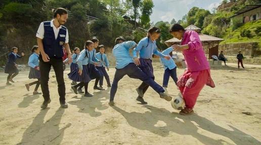 Children with blue shirt and blue pants and skirts playing football with Akira and their teacher in the school playground.