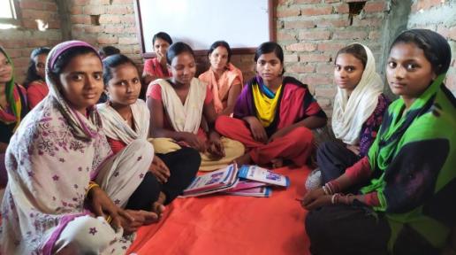 A group of eleven women wearing Kurthas are sitting on a red mattress inside a newly built room. There are notebooks in front of them and a whiteboard behind at the wall.