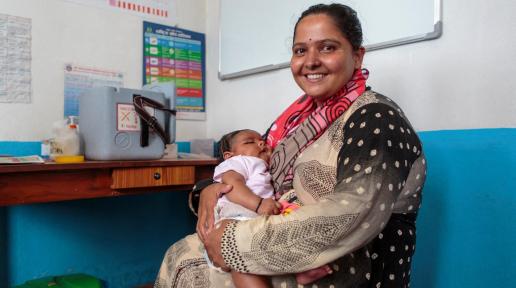 A mother holds her sleeping baby who's a few months old with a big smile on her face. The woman is sitting on a chair inside a room at the health post.