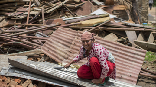 A woman wearing red kurtha and a scarf tied on her head looks at the camera with a smile as she examines the tin roof to rebuild a house.