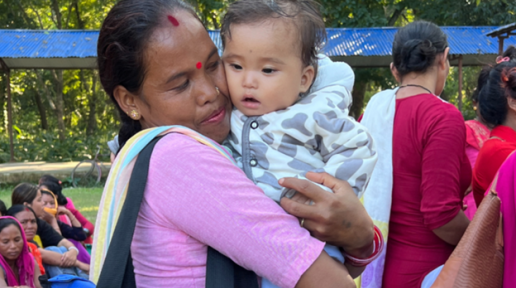A woman in a pink sari and a colorful scarf hugs a baby wearing a light blue cow-print puffer jacket. In the background there is a blue tin shed and other women.