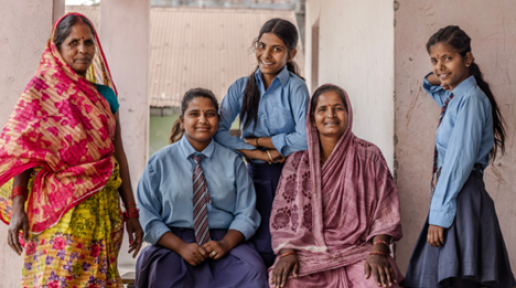 Three school girls wearing their school uniform blue shirt and blue skirt, are looking forward and smiling. There are two other middle aged women wearing sarees and smiling too.