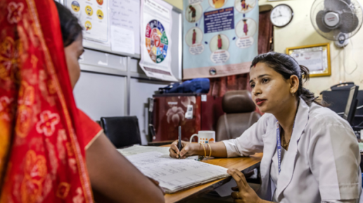 Psychosocial worker Kesh is wearing a white coat and intently listening to the woman in front of her wearing a red saree inside her office.