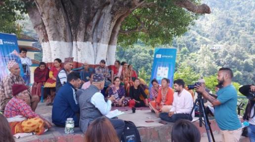 A group of locals sit on a seating area around a tree whilst journalists take pictures. Two blue UNESCO banners are positioned amongst them with trees in the background.