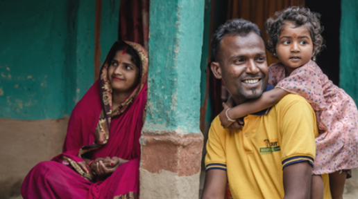 A woman wearing pink saree, red sindoor looks far ahead. Beside her is a man with a yellow t-shirt smiling and behind the man is an infant with a dress, hugging the man from behind.