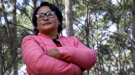 A women in a pink jacket and glasses, stands in the woods with her arms crisscrossed gazing into the distance.