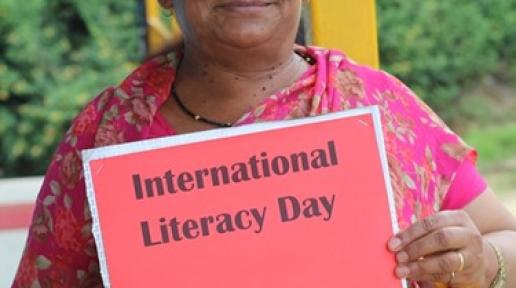 Chhalmaya wears a pink Saree with flower designs, holds a red paper that says International Literacy Day and smiles looking forward.