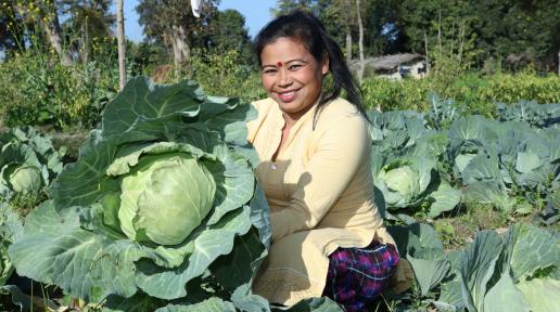 A woman stands in the field with a joyful expression, proudly holding a large cabbage she has harvested.