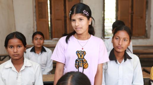 A young girl is standing in a classroom, surrounded by her friends who are seated on benches.